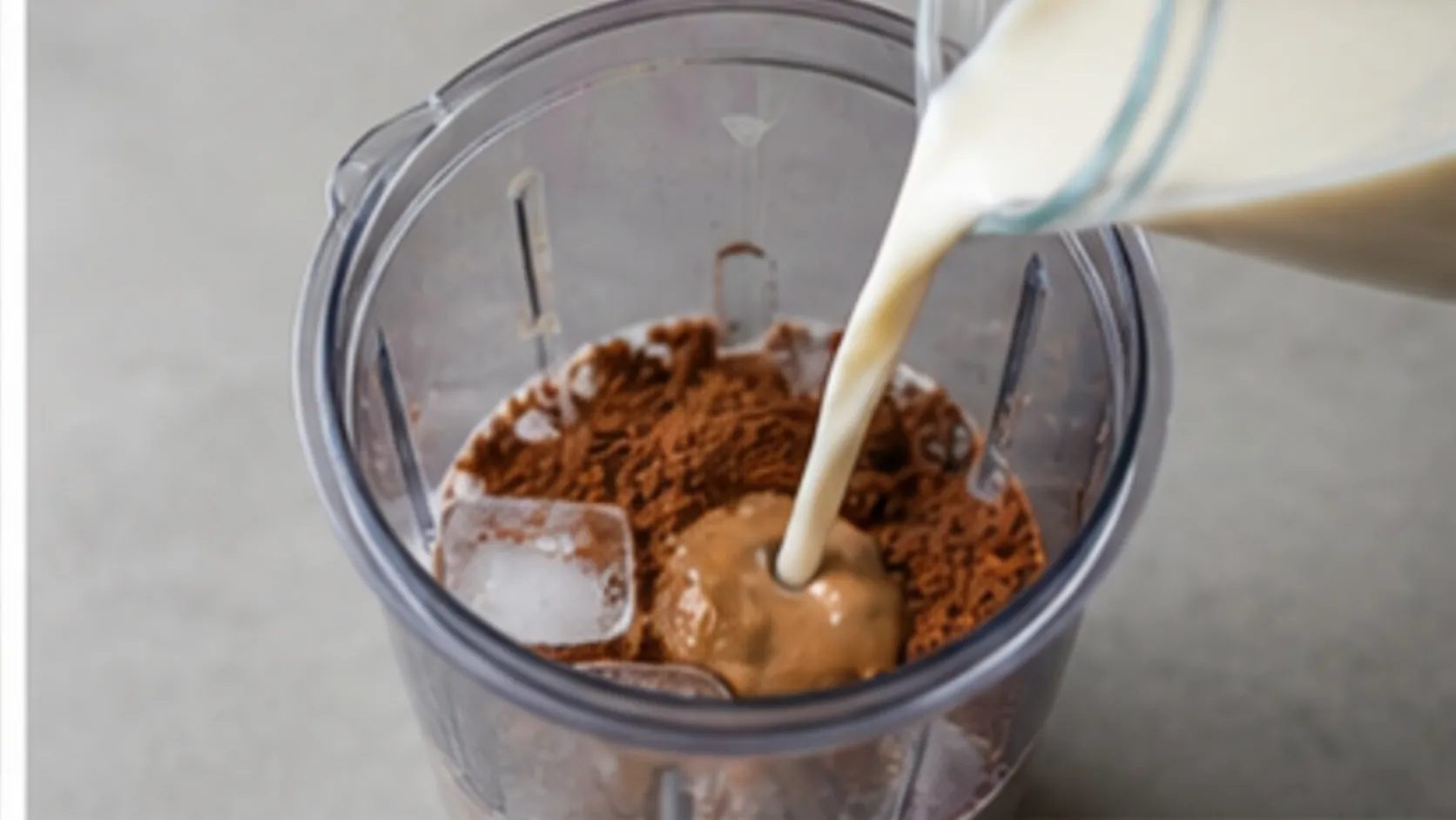 Almond milk being poured into a blender with almond butter, baking cocoa and ice cubes